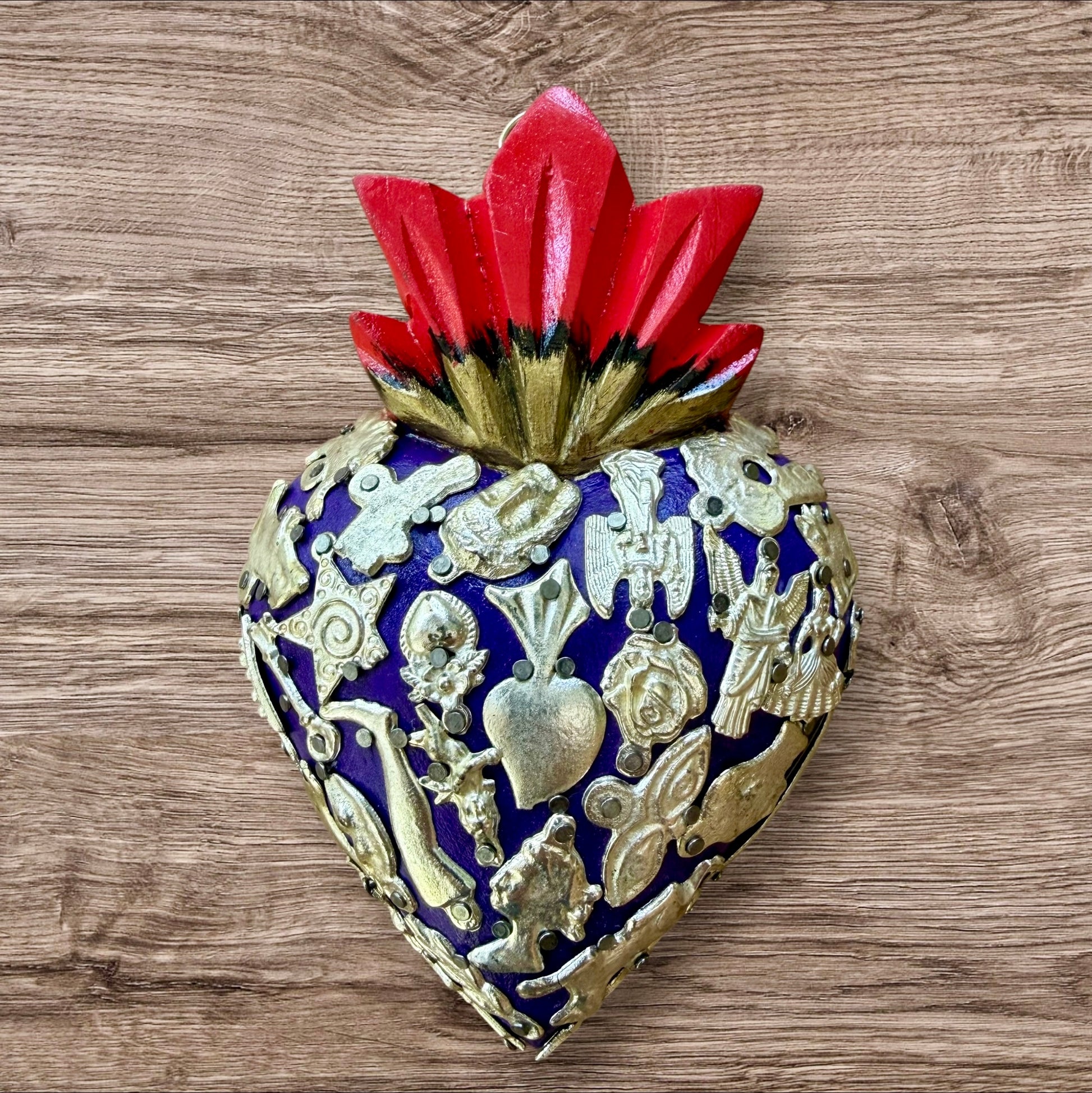 A Milagros heart featuring a gold textured top and a purple base covered in raised silver-toned charms displayed on a wooden wall.