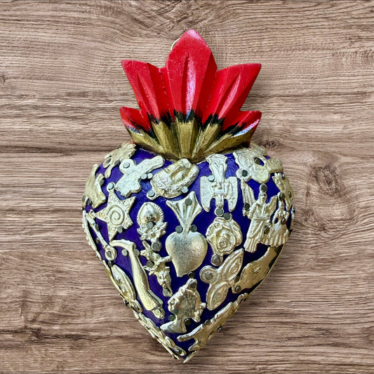 A Milagros heart featuring a gold textured top and a purple base covered in raised silver-toned charms displayed on a wooden wall.