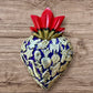 A Milagros heart featuring a gold textured top and a purple base covered in raised silver-toned charms displayed on a wooden wall.