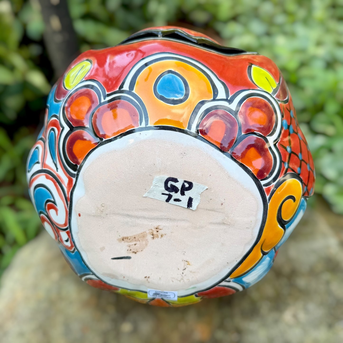 A hand-painted ceramic jack-o'-lantern pumpkin with a red face, black carved eyes and mouth, and a multicolored floral pattern on a garden background, bottom view. 