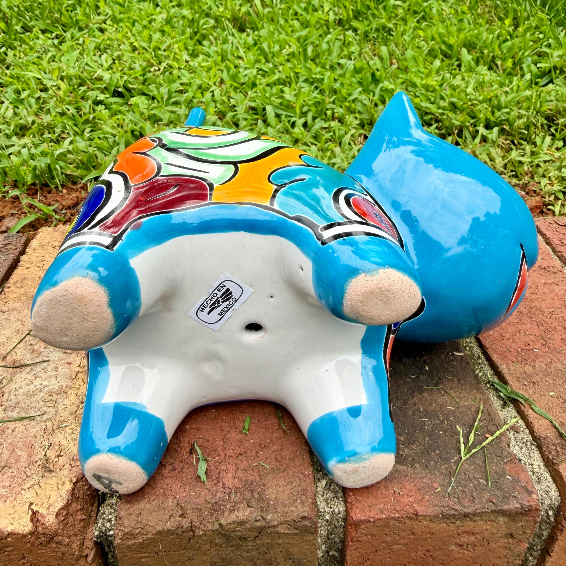 A glossy talavera ceramic cat figurine decorated with vibrant hand-painted patterns in turquoise, yellow, green and red, standing on a brick path with a green lawn and a house in the blurred background. View of the figurine bottom.