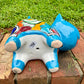 A glossy talavera ceramic cat figurine decorated with vibrant hand-painted patterns in turquoise, yellow, green and red, standing on a brick path with a green lawn and a house in the blurred background. View of the figurine bottom.