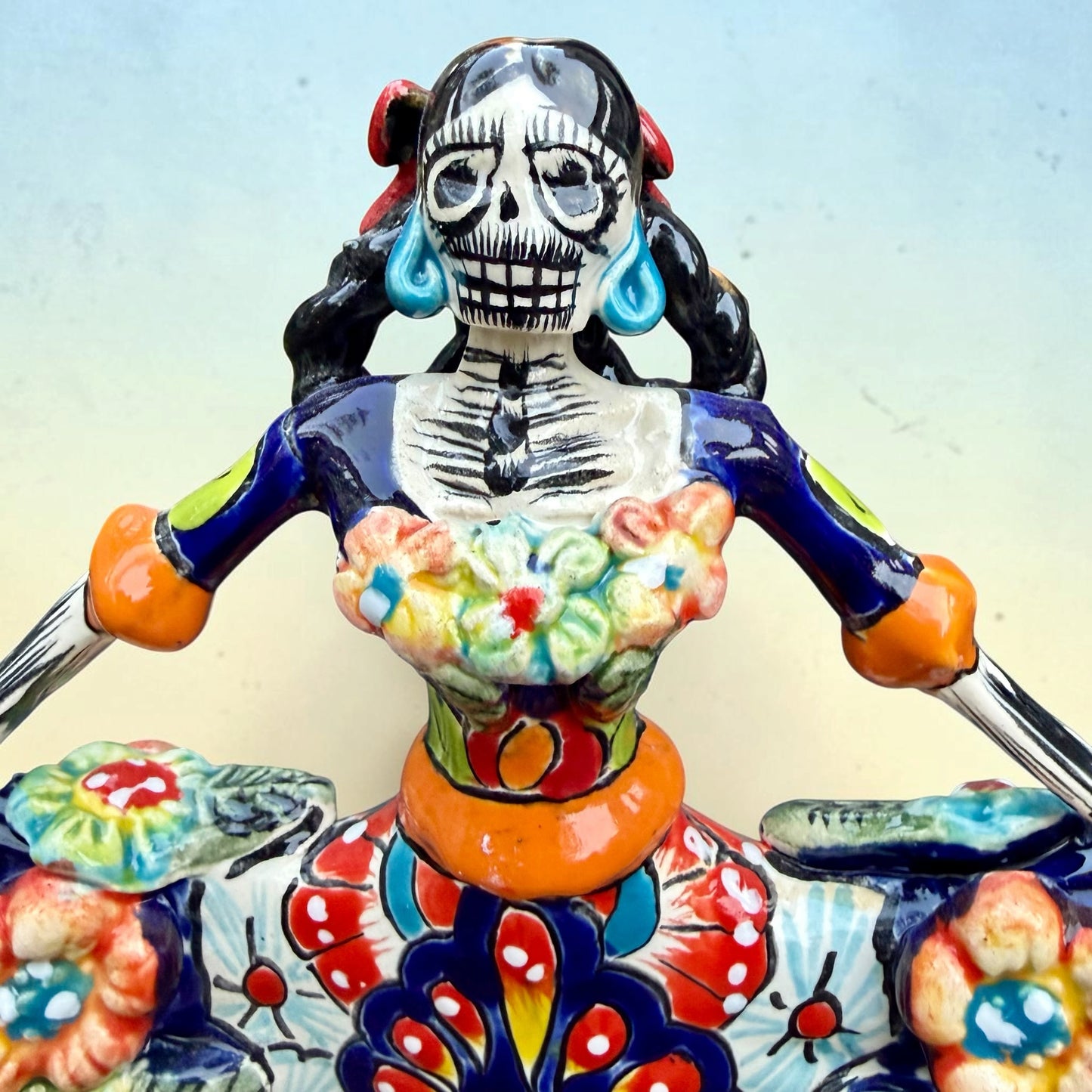 A colorful talavera Day of the Dead (Día de los Muertos) dancing skeleton ballerina figurine. The figurine features black hair in braids wearing a vibrant multicolor dress with floral patterns and orange accents, set against a light blue and beige gradient background. This is a close up of her face.