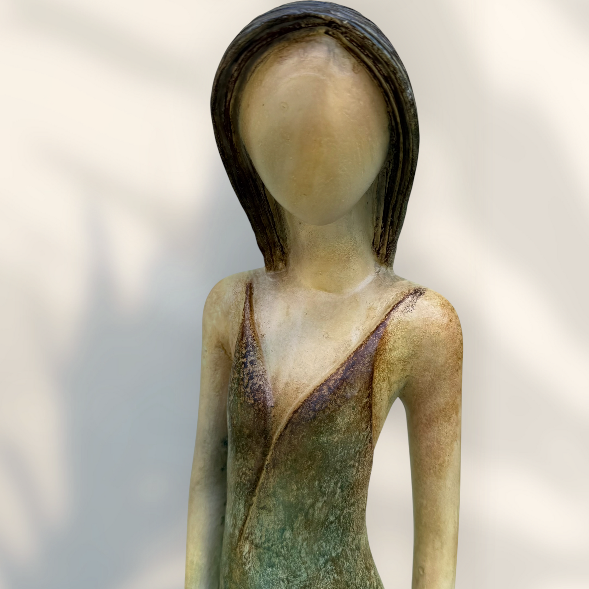 A Loretta Bertoli statue of a faceless woman in a long flowing dress with two hummingbirds, displayed against a stacked stone tile wall on a light tiled floor. Face close up.