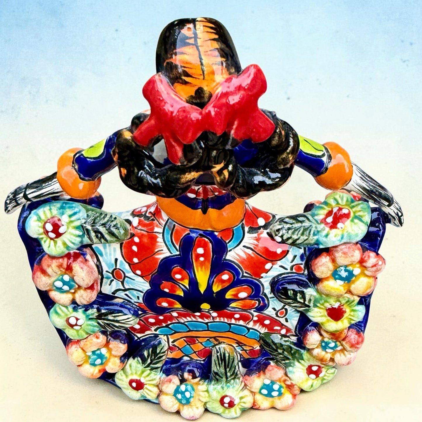 A colorful talavera Day of the Dead (Día de los Muertos) dancing skeleton ballerina figurine. The figurine features black hair in braids wearing a vibrant multicolor dress with floral patterns and orange accents, set against a light blue and beige gradient background, top view from back.