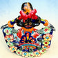A colorful talavera Day of the Dead (Día de los Muertos) dancing skeleton ballerina figurine. The figurine features black hair in braids wearing a vibrant multicolor dress with floral patterns and orange accents, set against a light blue and beige gradient background, top view from back.