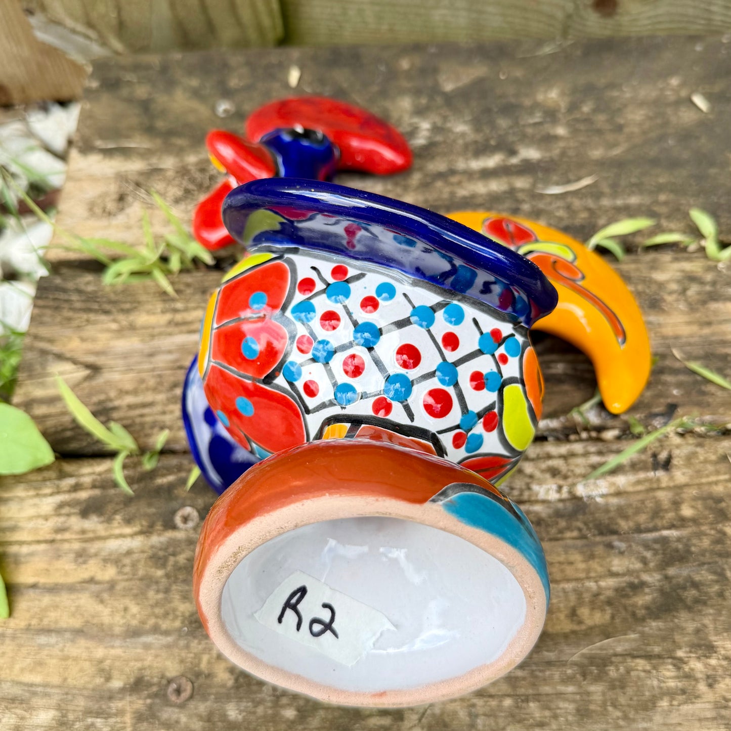 A colorful ceramic rooster figurine standing on a weathered wooden surface outdoors, with a green wooden wall behind it. The rooster is decorated with bright red, yellow, blue, and orange painted details, bottom of Rooster view.