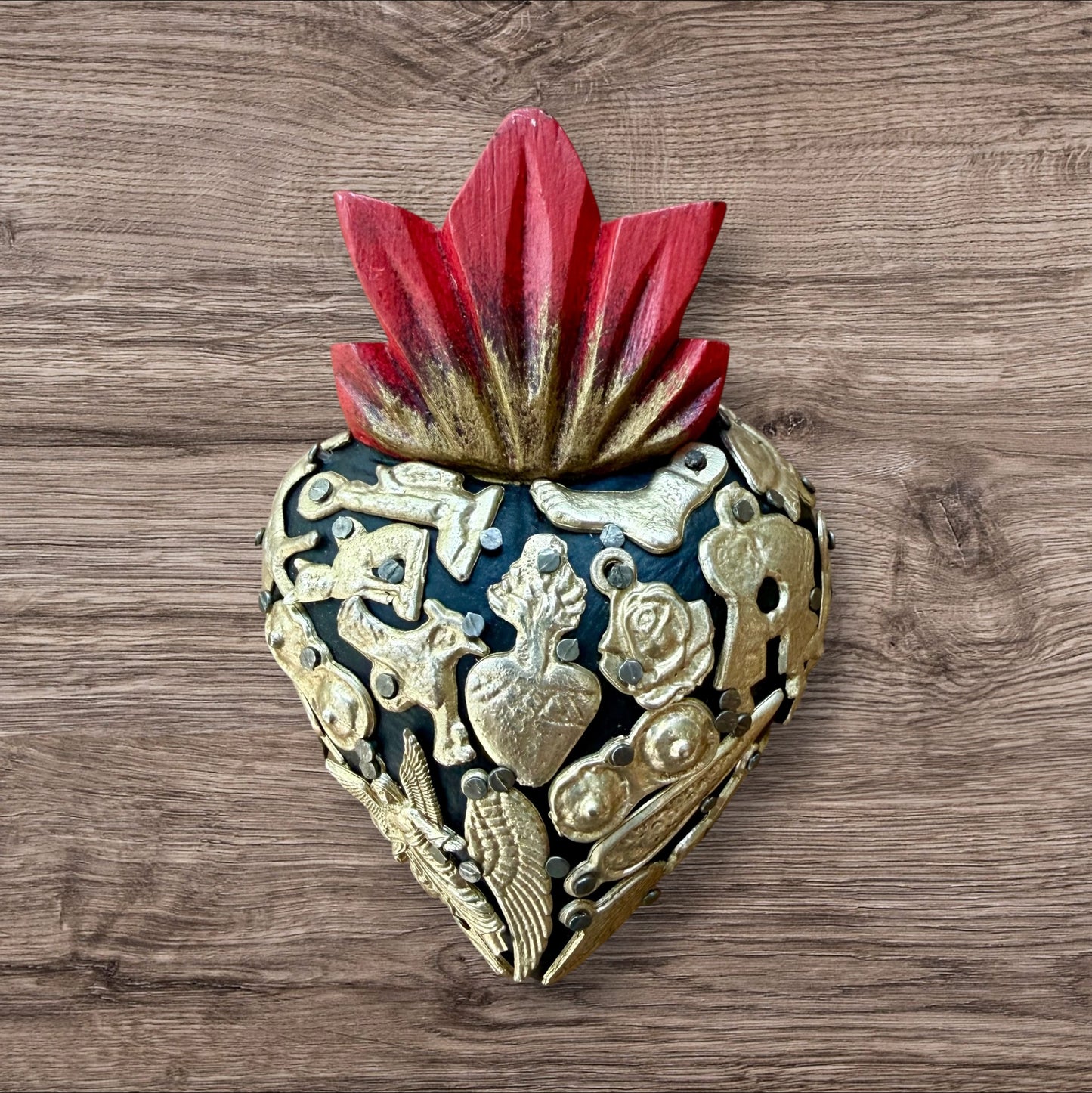 A Mexican Milagros heart in black with a gold flaming red top, covered in golden charms. 