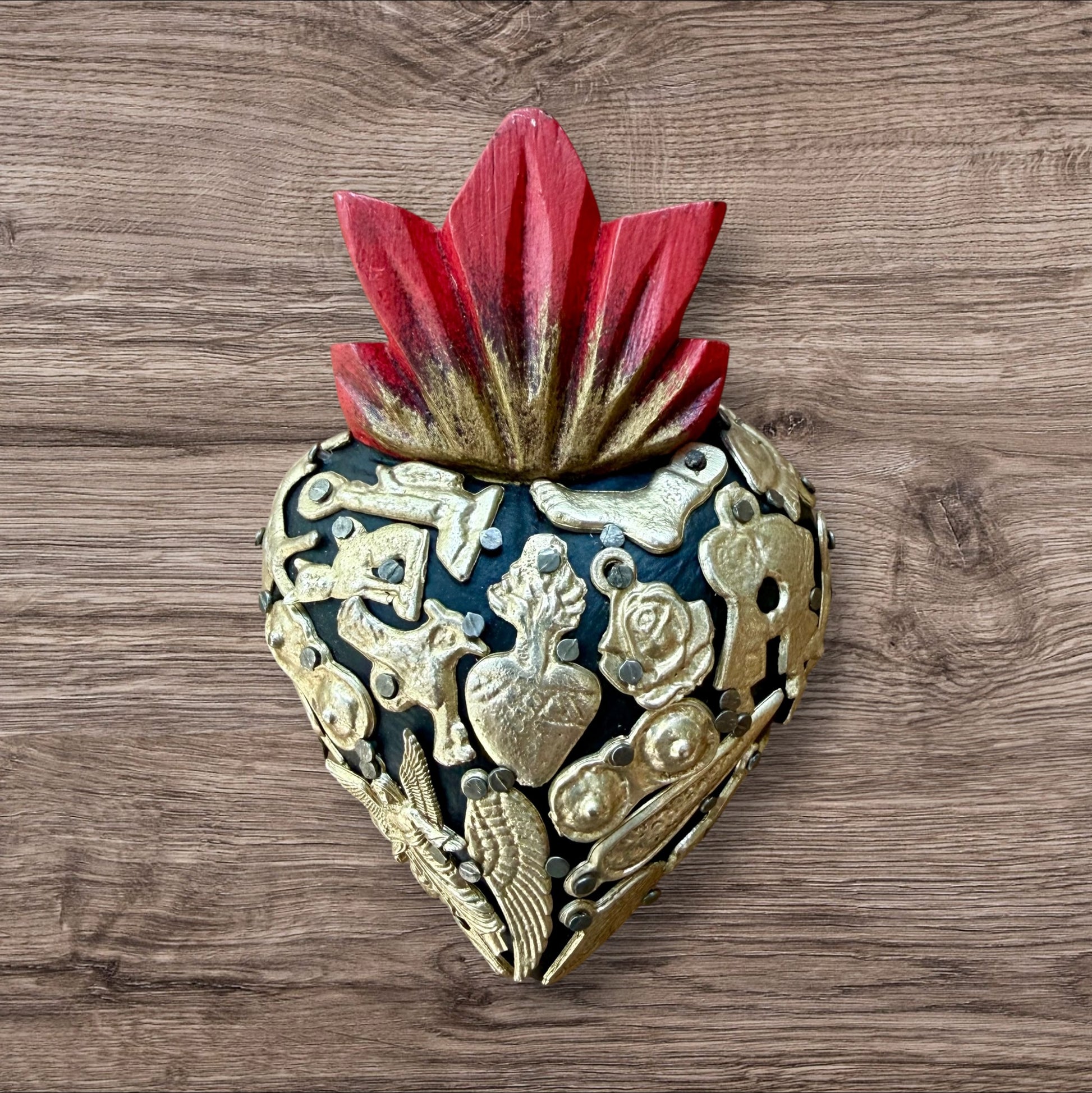 A Mexican Milagros heart in black with a gold flaming red top, covered in golden charms. 