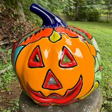 A hand-painted ceramic jack-o'-lantern pumpkin ornament with a bright orange face, red carved eyes and mouth, a blue stem, and colorful side patterns, sitting outdoors on a stone pedestal in a grassy yard.