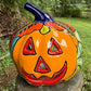A hand-painted ceramic jack-o'-lantern pumpkin ornament with a bright orange face, red carved eyes and mouth, a blue stem, and colorful side patterns, sitting outdoors on a stone pedestal in a grassy yard.