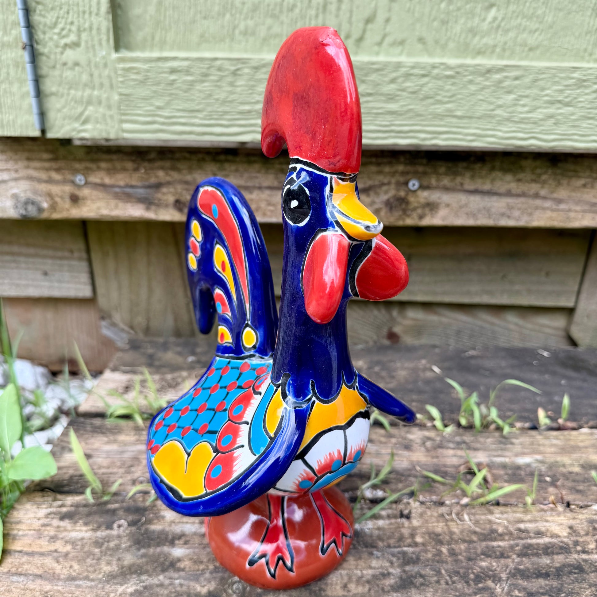 A colorful hand-painted ceramic rooster figurine standing on a rustic wooden surface in front of a green wooden door.