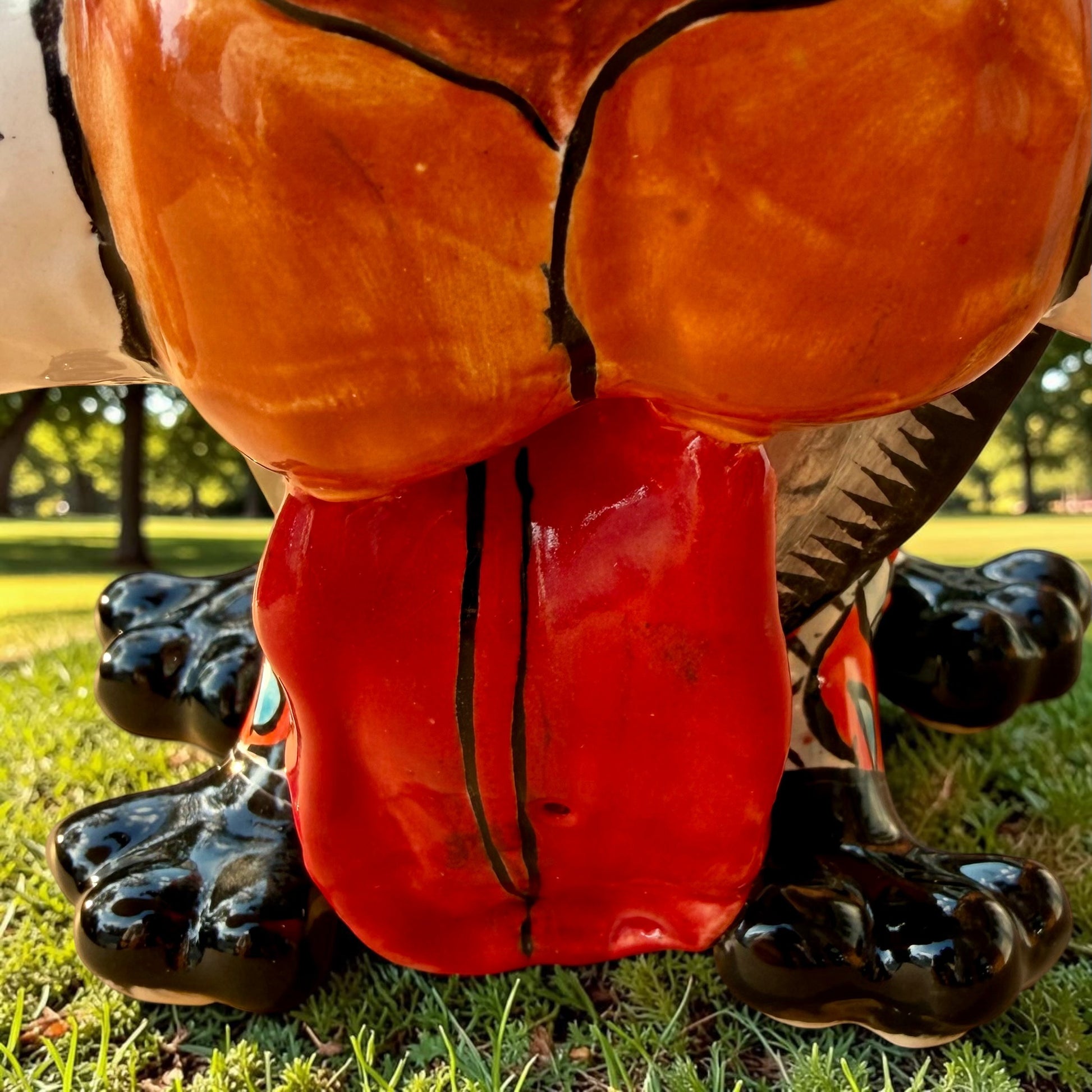 A colorful Talavera dog statue holding a big white bone, photographed outdoors on grass with trees in the background. The dog has a glossy black body, blue eyes, an orange snout, and multicolored patterned ears.  Close up of his red tongue .
