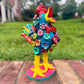 A colorful rooster folk art sculpture with a blue body, yellow legs and beak, and many bright flower ornaments, standing on a pink circular base outdoors on a brick path with a blurred green lawn behind it. Left angle view of Rooster.