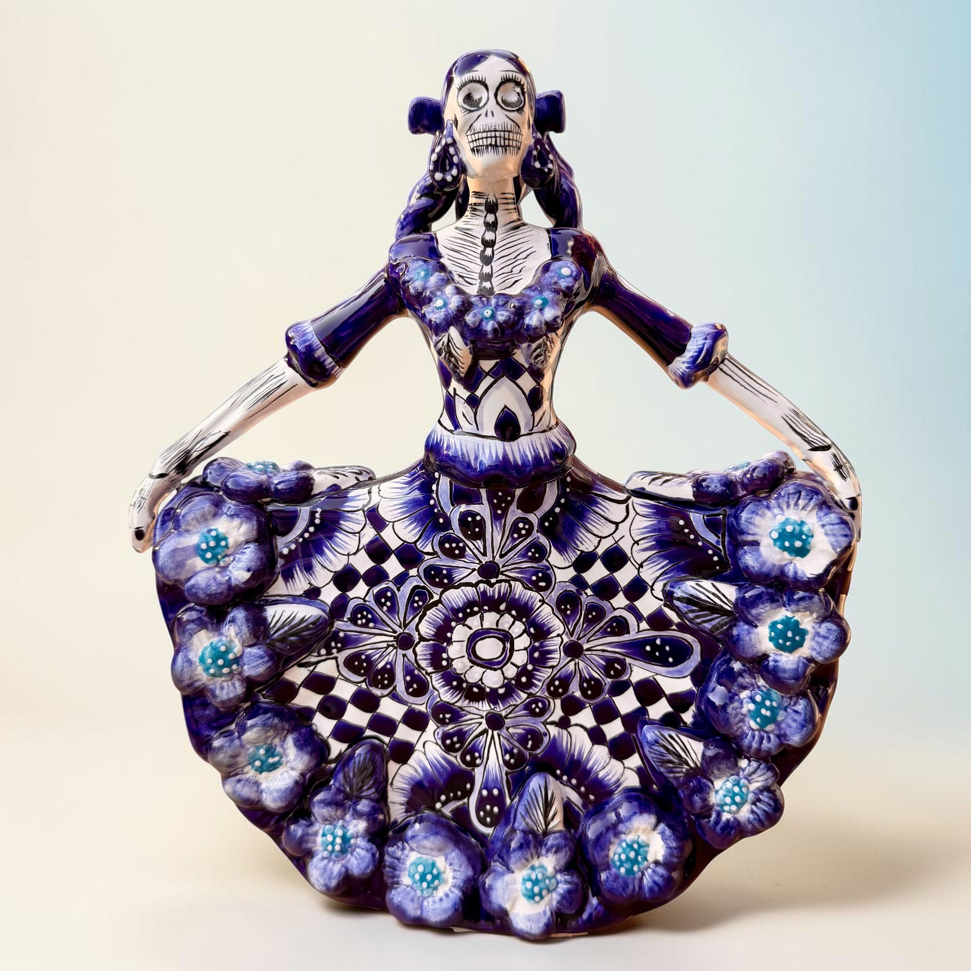 A Day of the Dead style talavera figurine of a skeleton woman in a wide floral skirt, painted in deep blue, black, white, and light blue on a soft beige-to-blue gradient background.