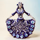 A Day of the Dead style talavera figurine of a skeleton woman in a wide floral skirt, painted in deep blue, black, white, and light blue on a soft beige-to-blue gradient background.