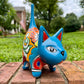 A glossy talavera ceramic cat figurine decorated with vibrant hand-painted patterns in turquoise, yellow, green and red, standing on a brick path with a green lawn and a house in the blurred background.