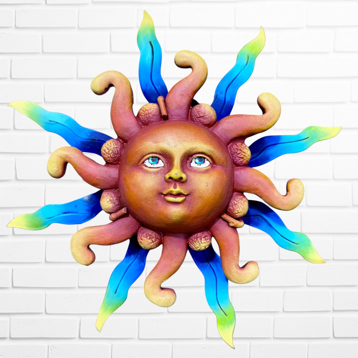 A whimsical sun wall sculpture with big blue eyes, a smiling face, and vibrant blue rays mounted on a white brick wall.
