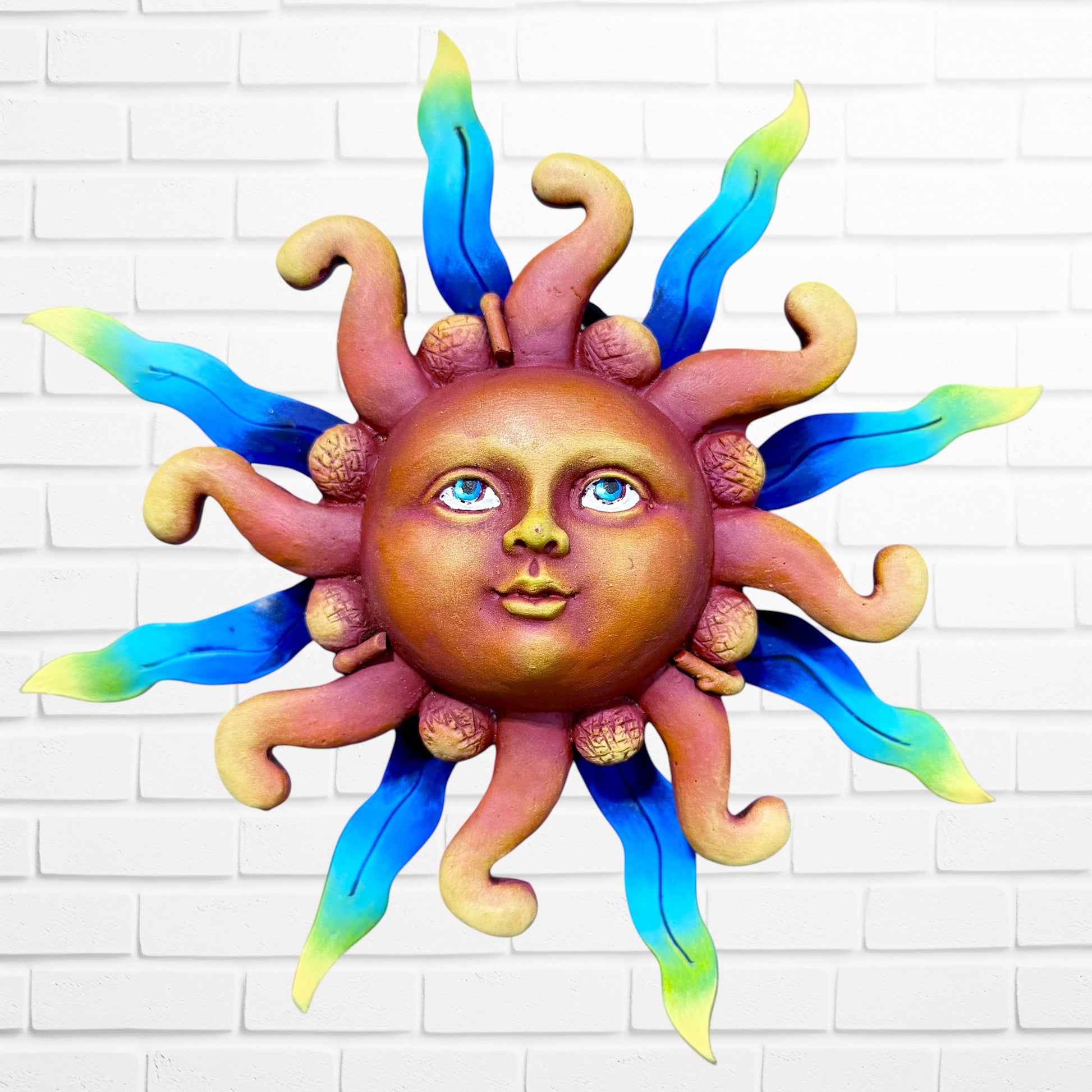 A whimsical sun wall sculpture with big blue eyes, a smiling face, and vibrant blue rays mounted on a white brick wall.