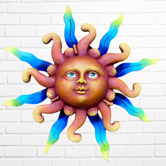 A whimsical sun wall sculpture with big blue eyes, a smiling face, and vibrant blue rays mounted on a white brick wall.