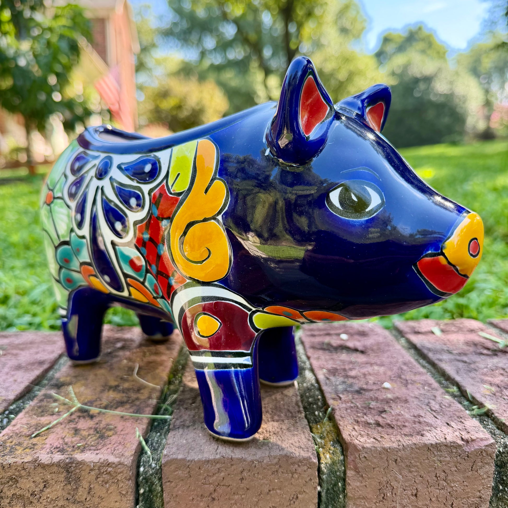 A glossy talavera pig figurine decorated with vibrant hand-painted patterns in blue, yellow, red, green, and white, standing on a brick surface with a blurred outdoor garden background.