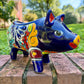 A glossy talavera pig figurine decorated with vibrant hand-painted patterns in blue, yellow, red, green, and white, standing on a brick surface with a blurred outdoor garden background.