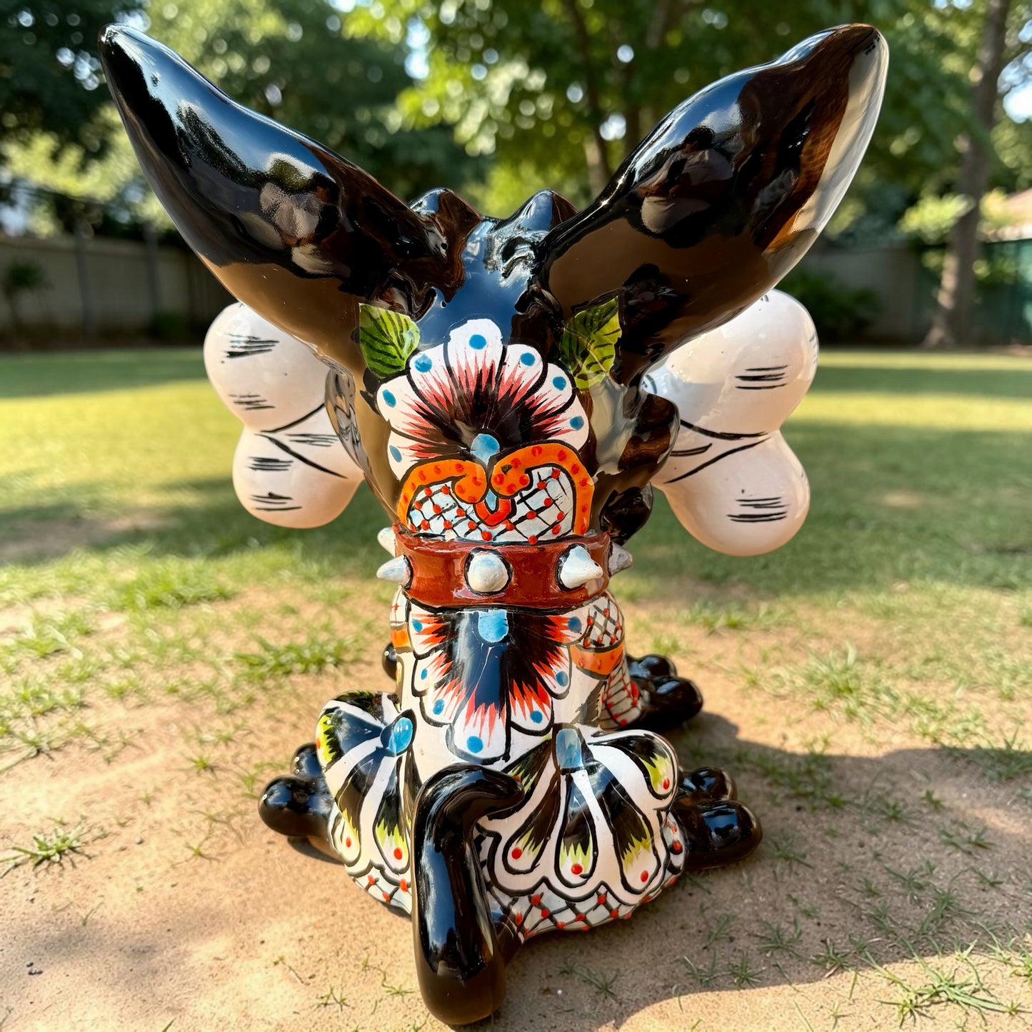 A colorful Talavera dog statue holding a big white bone, photographed outdoors on grass with trees in the background. The dog has a glossy black body, blue eyes, an orange snout, and multicolored patterned ears.  Back view of the dog showing his curled tail.