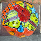 A hand-painted Talavera  jack-o'-lantern pumpkin sits on a brick patio with a green grass lawn and trees in the background. The pumpkin features a glossy orange base with hand-painted details in red, yellow, blue, and green. Top view of pumpkin showing stem.