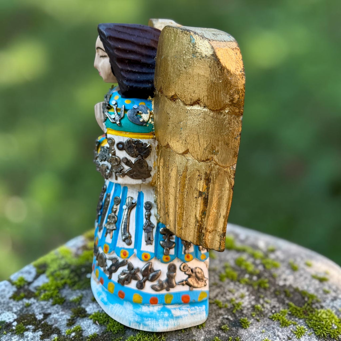 A Milagros guardian angel figurine with gold wings, dark hair, and a blue, white, and gold embroidered dress, posed outdoors on a stone surface with a blurred green background. This is the right side view.