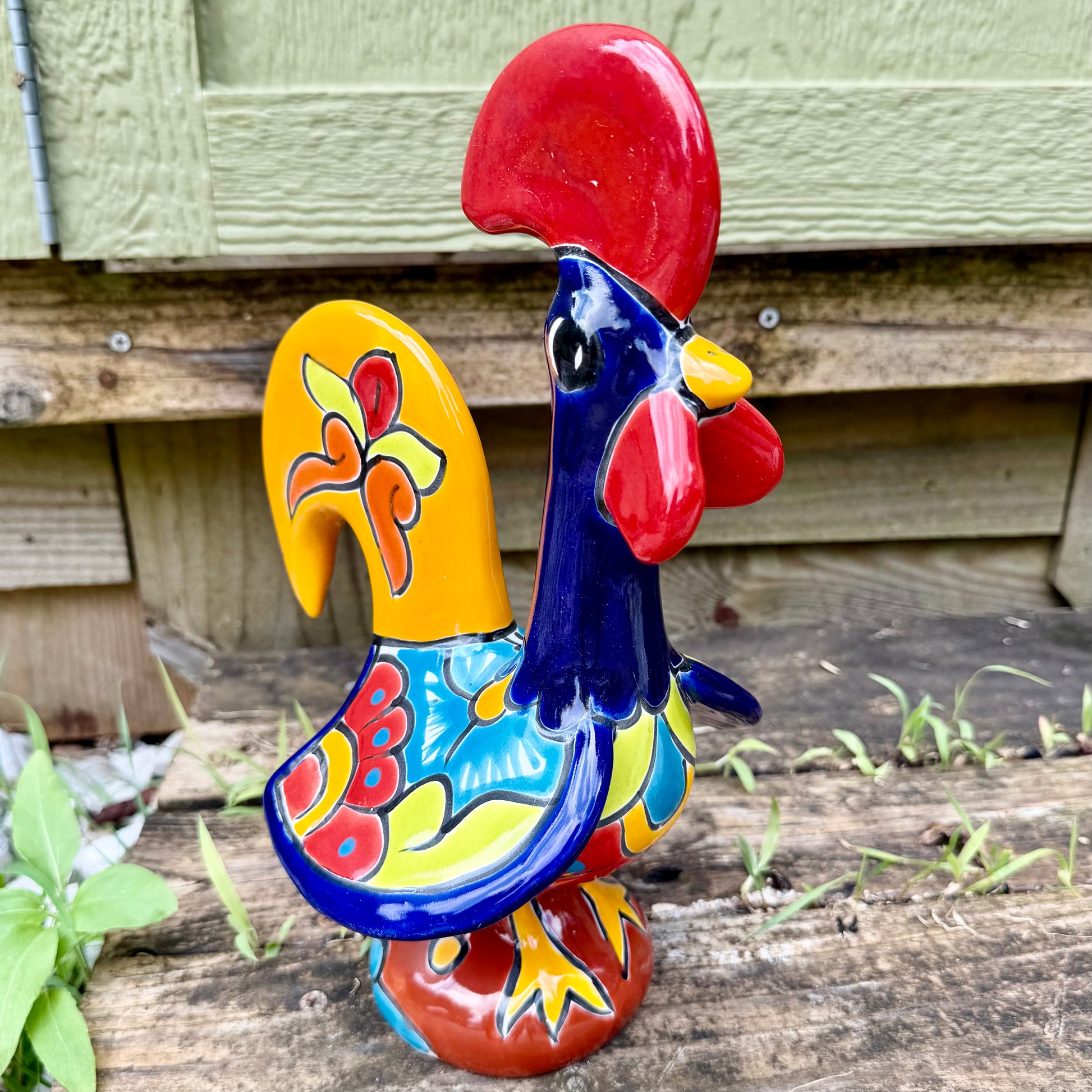 A colorful ceramic rooster figurine standing on a weathered wooden surface outdoors, with a green wooden wall behind it. The rooster is decorated with bright red, yellow, blue, and orange painted details.
