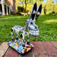 A colorful ceramic donkey Catrina figurine with a skeletal white-and-black design and a floral base, photographed outdoors on a brick surface with a blurred green lawn and house in the background.