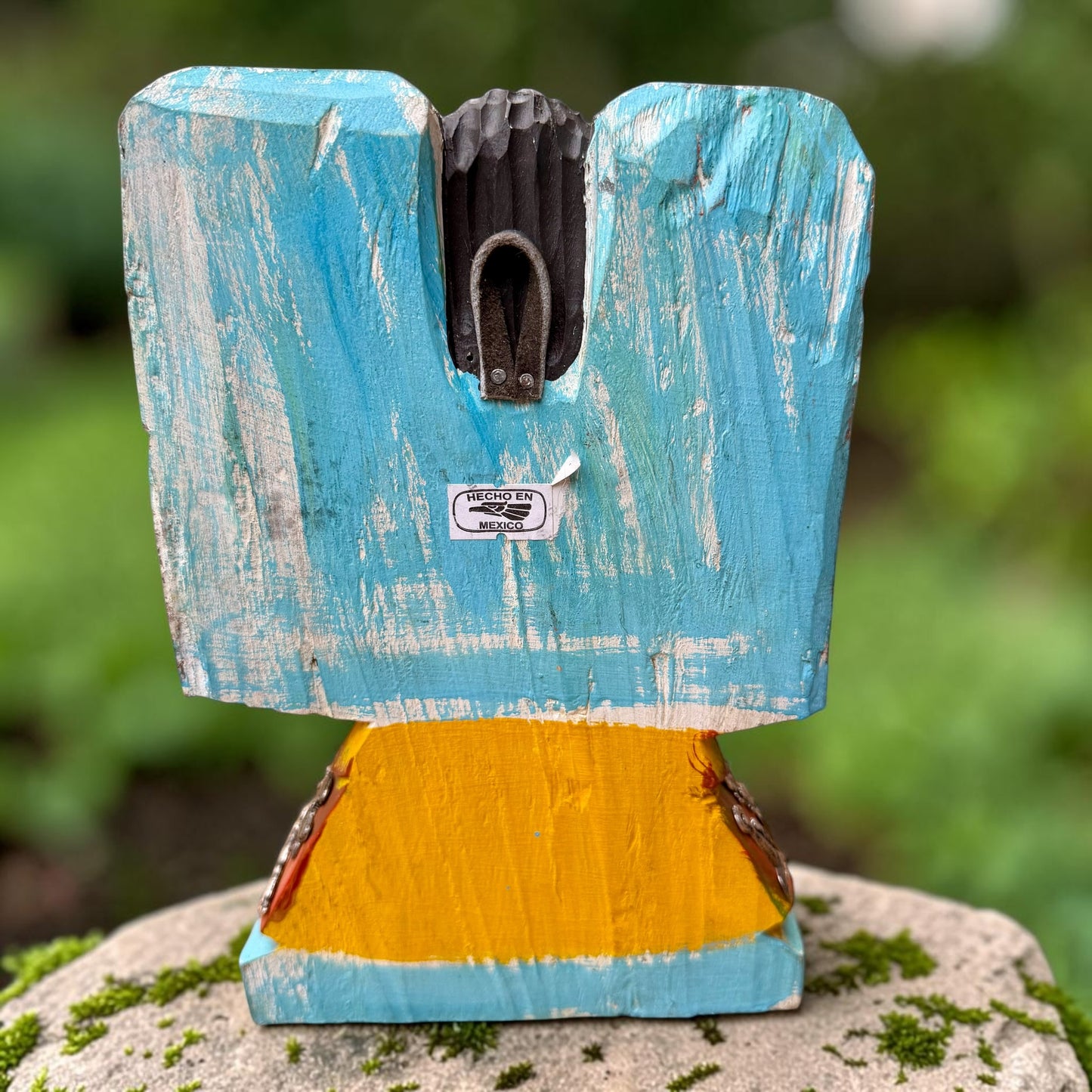 A handcrafted wooden Milagros angel figurine with large wings, decorated with metallic silver charms and painted in yellow, and orange, standing on a mossy stone with a blurred green garden background. Back view of angle showing hanging loop