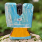 A handcrafted wooden Milagros angel figurine with large wings, decorated with metallic silver charms and painted in yellow, and orange, standing on a mossy stone with a blurred green garden background. Back view of angle showing hanging loop