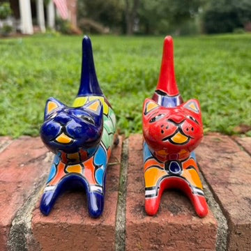 Two colorful ceramic cat figurines, one blue and one red, decorated with geometric patterns, displayed on a brick ledge with a green lawn in the background front face view.