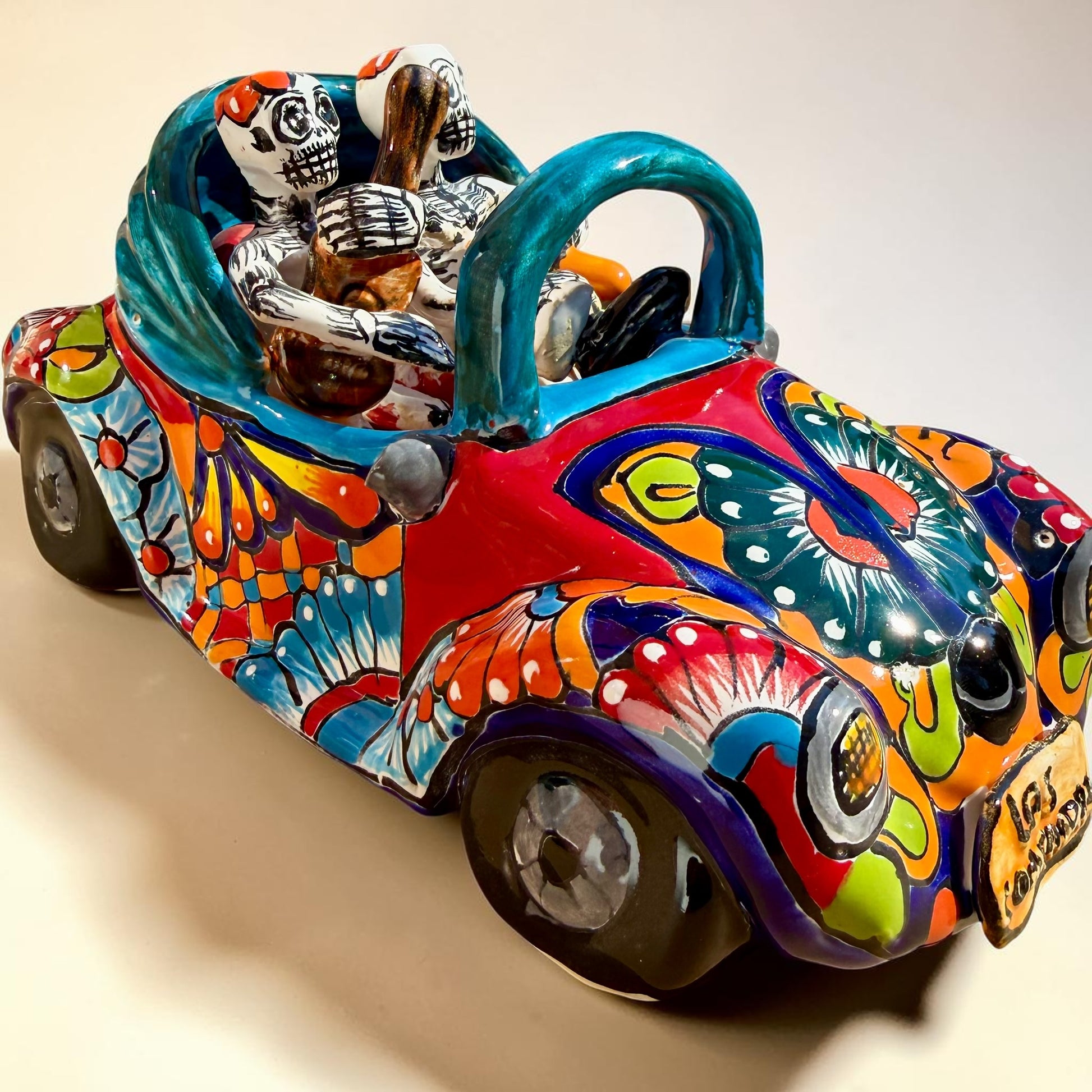 A colorful hand-painted ceramic Mexican-style convertible car figurine with Day of the Dead skeleton characters inside, shown on a neutral beige background.