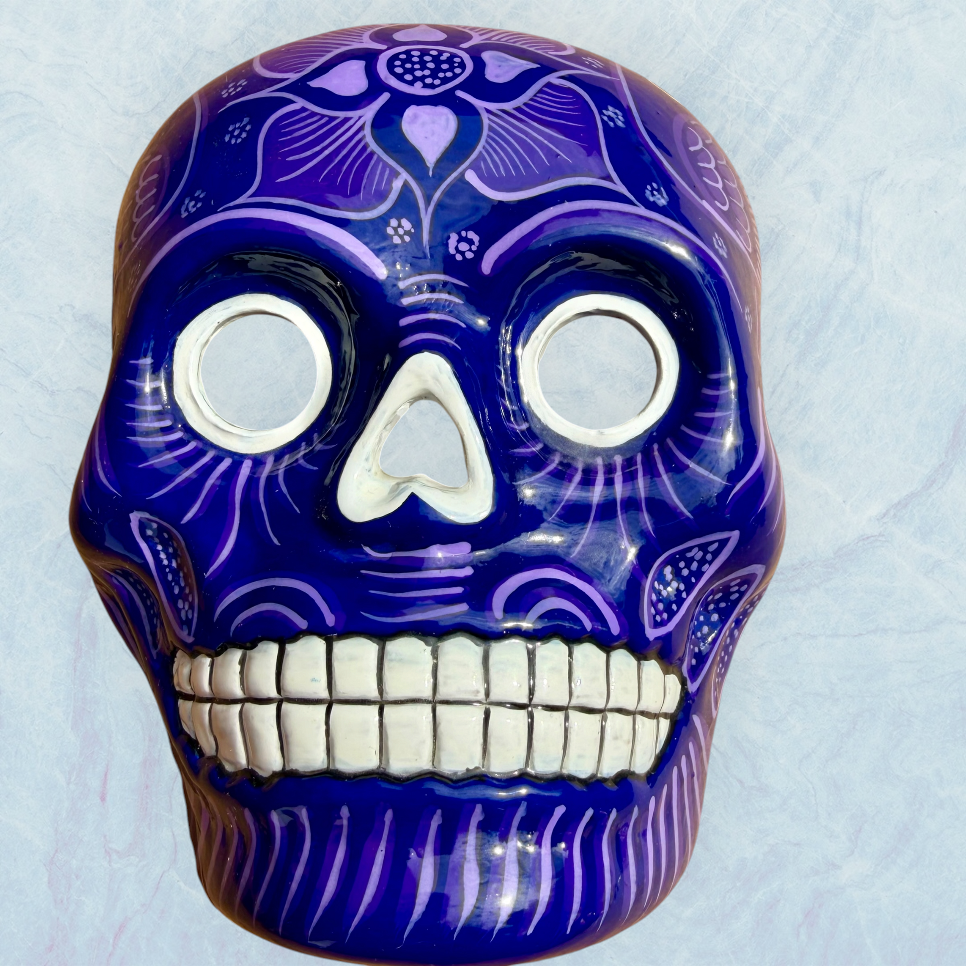 A clay, purple with designs, Mexican-style skeleton skull face mask hanging on a soft blue surface.
