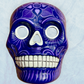 A clay, purple with designs, Mexican-style skeleton skull face mask hanging on a soft blue surface.