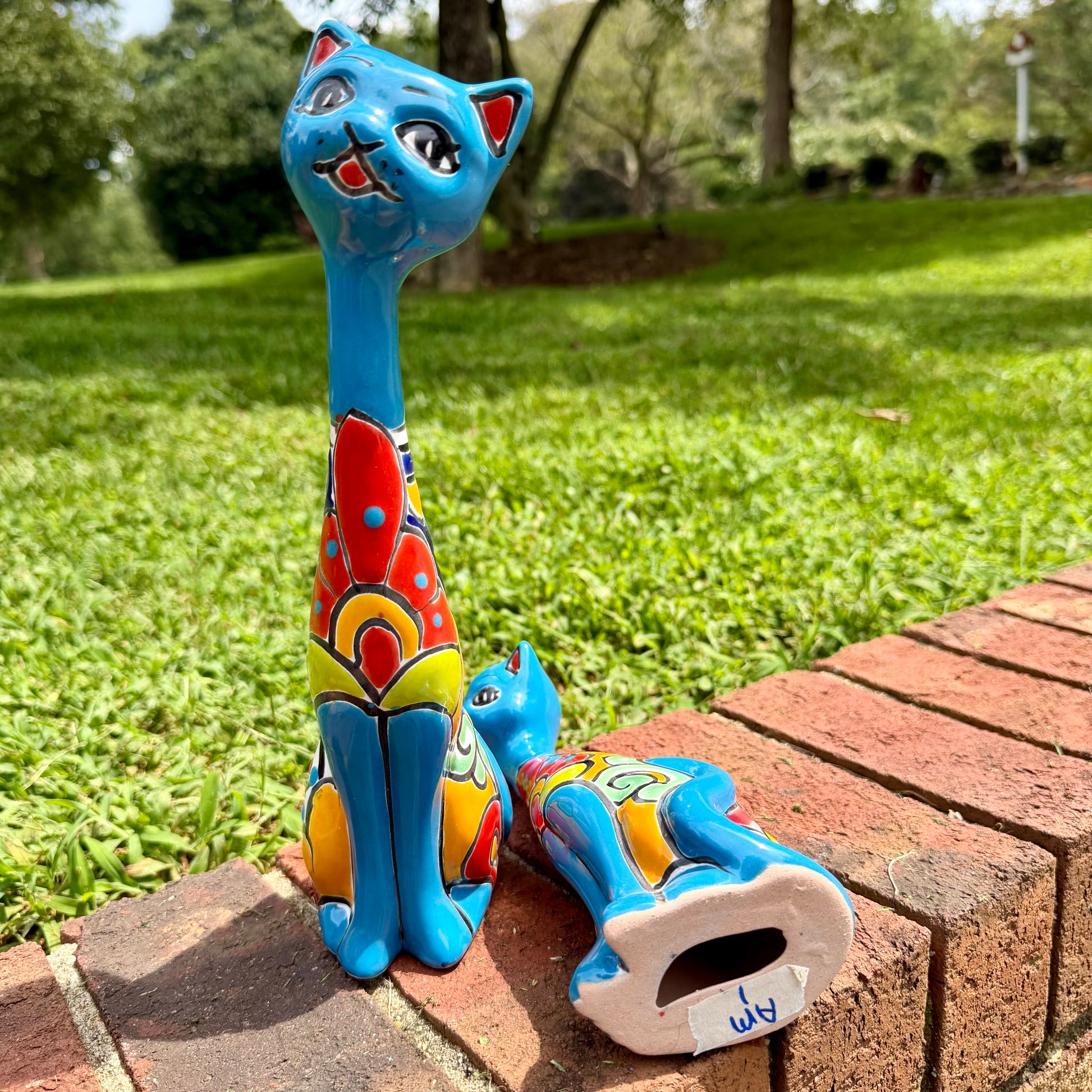 A colorful hand-painted ceramic cat figurine stands on a brick ledge with a blurred green lawn and trees in the background. A second matching figurine lies on its side nearby.