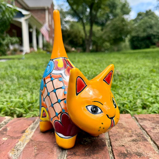 A hand-painted ceramic cat figurine in bright yellow with red, blue, and beige patterns, standing on a brick surface with a blurred green lawn and house in the background.