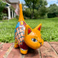 A hand-painted ceramic cat figurine in bright yellow with red, blue, and beige patterns, standing on a brick surface with a blurred green lawn and house in the background.