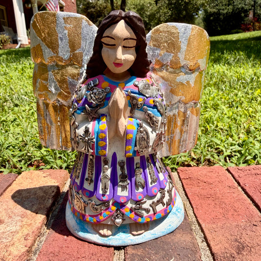 A colorful painted wood angel with milagros with silver and gold wings and a decorated robe, standing outdoors on red bricks in a grassy yard.