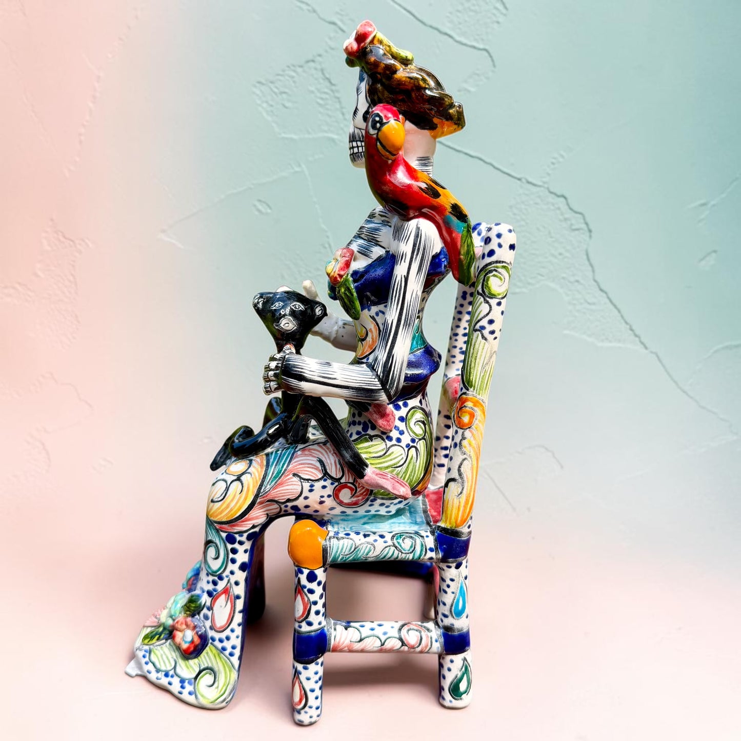 Frida Kahlo Talavera day of the dead figurine sitting in a chair holding a monkey with a parrot on her shoulder, left side view.
