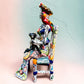 Frida Kahlo Talavera day of the dead figurine sitting in a chair holding a monkey with a parrot on her shoulder, left side view.