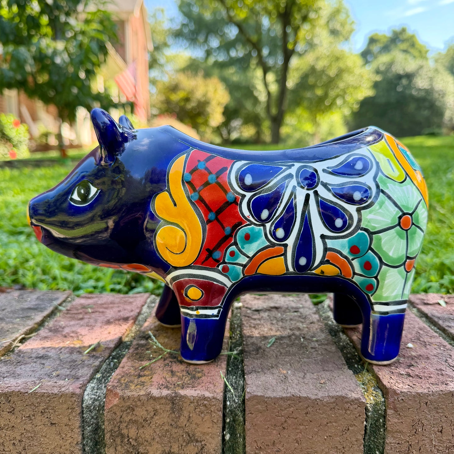 A glossy talavera pig figurine decorated with vibrant hand-painted patterns in blue, yellow, red, green, and white, standing on a brick surface with a blurred outdoor garden background, right side view.