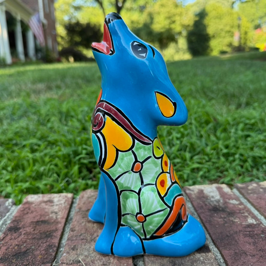 A hand-painted talavera ceramic dog figurine in a sitting position, featuring a vibrant blue base with colorful geometric patterns in red, yellow, orange, and white. The figurine is photographed outdoors on a brick surface with a blurred green lawn and trees in the background side view