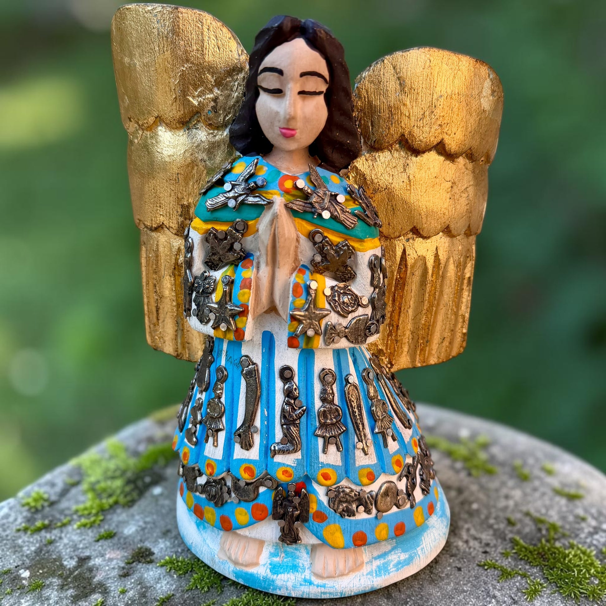 A Milagros guardian angel figurine with gold wings, dark hair, and a blue, white, and gold embroidered dress, posed outdoors on a stone surface with a blurred green background.