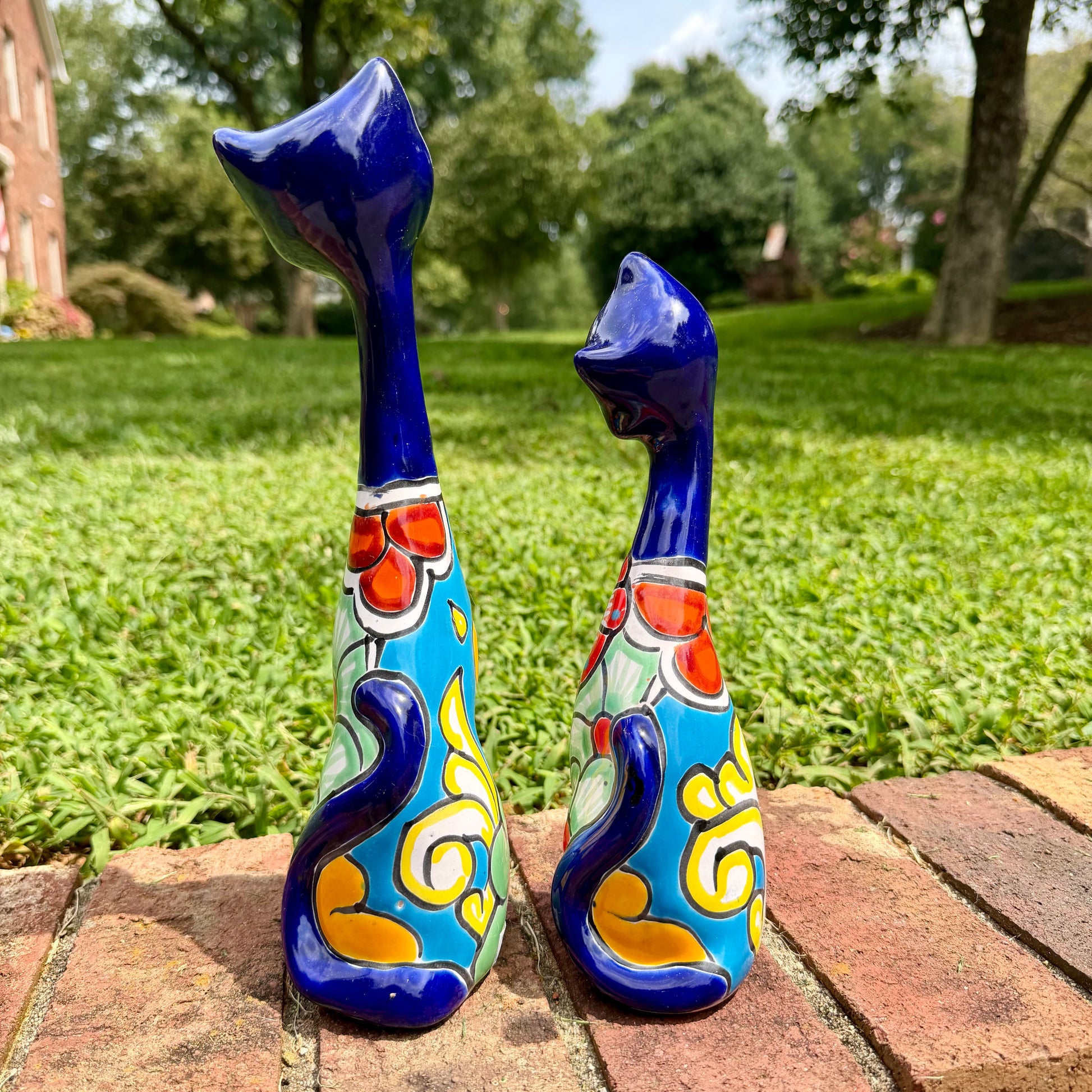 Two decorative ceramic cat figurines with long blue necks and colorful floral patterns, standing on a brick path in a grassy outdoor garden setting. this is the view of their backs