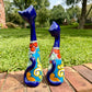 Two decorative ceramic cat figurines with long blue necks and colorful floral patterns, standing on a brick path in a grassy outdoor garden setting. this is the view of their backs