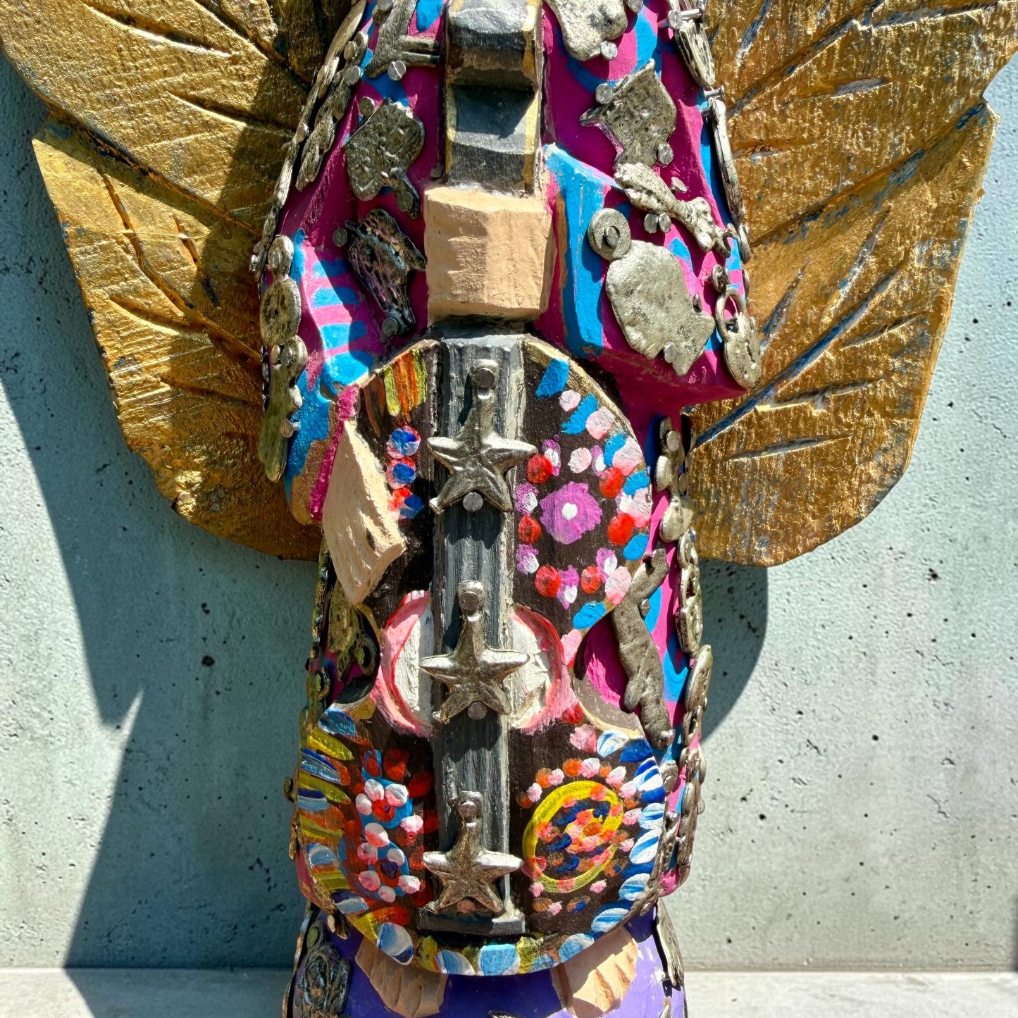 A handmade wood Milagros angel figurine with large gold wings, holding a guitar, dressed in a colorful patterned robe against a textured blue wall, guitar close up.