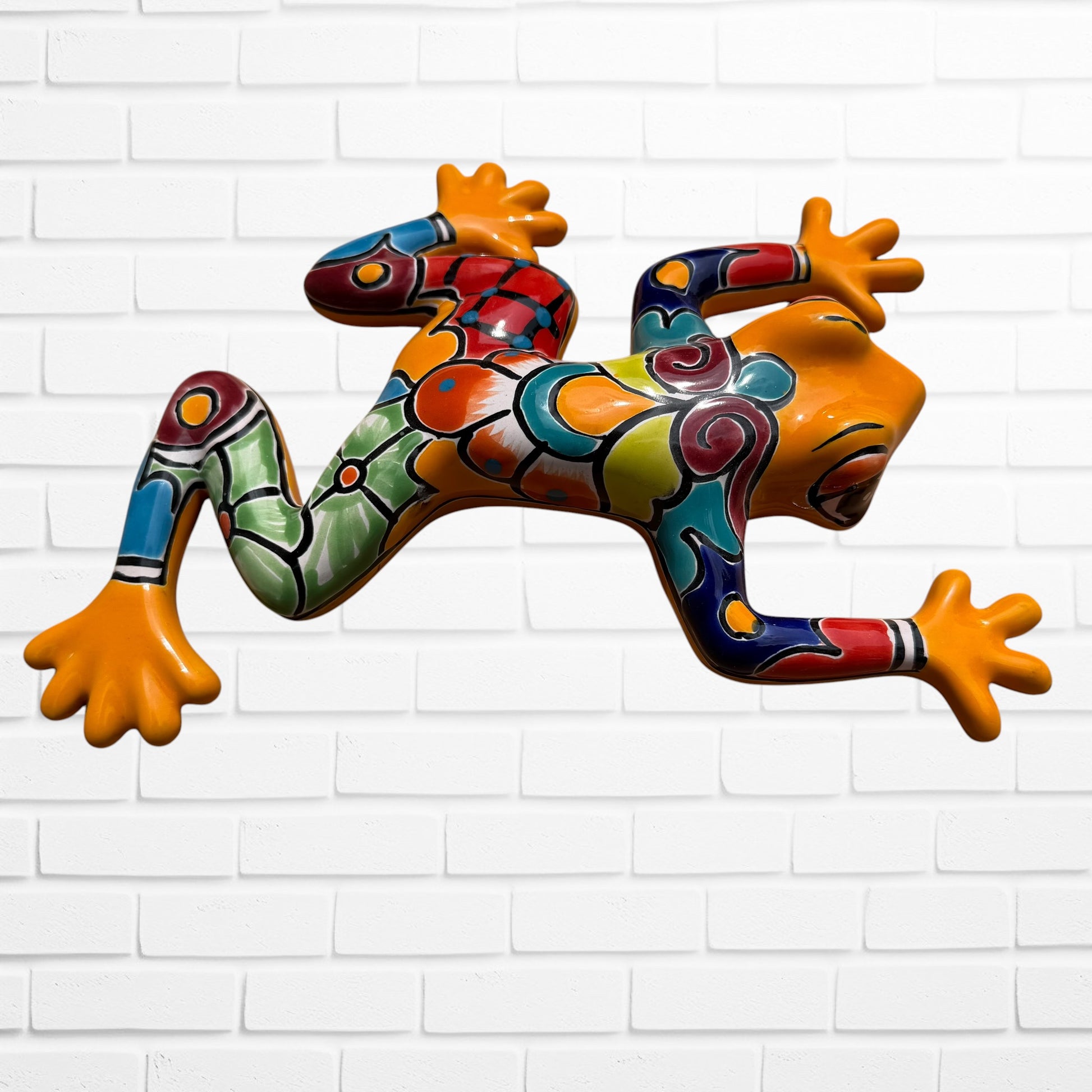 A colorful, glossy ceramic frog sculpture mounted on a white brick wall. The frog features a vibrant, multi-colored pattern with orange, green, blue, red, and yellow sections separated by black outlines.